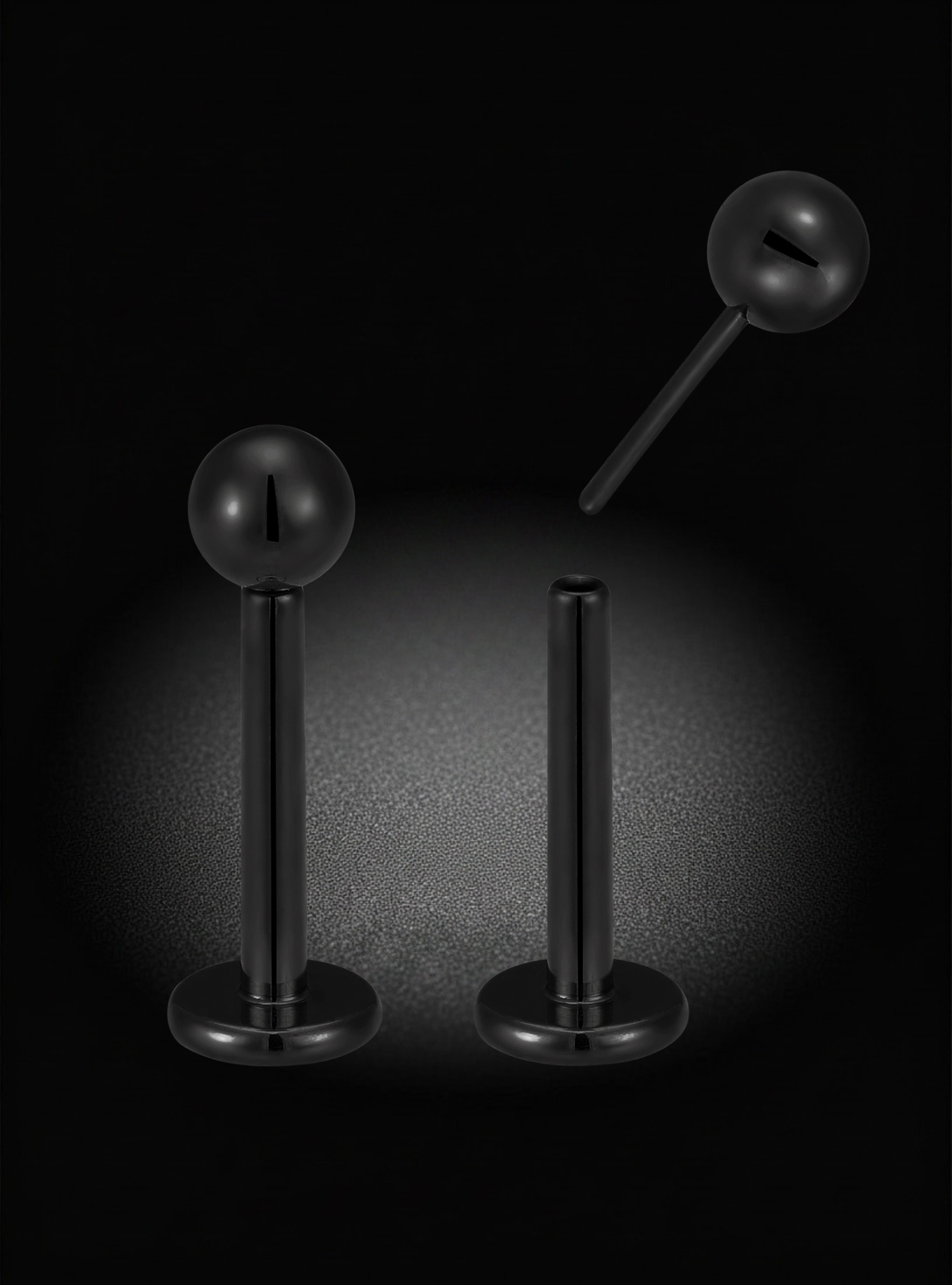 Three black labret-style body jewelry piercings with spherical ball ends are displayed against a dark background with a spotlight effect.