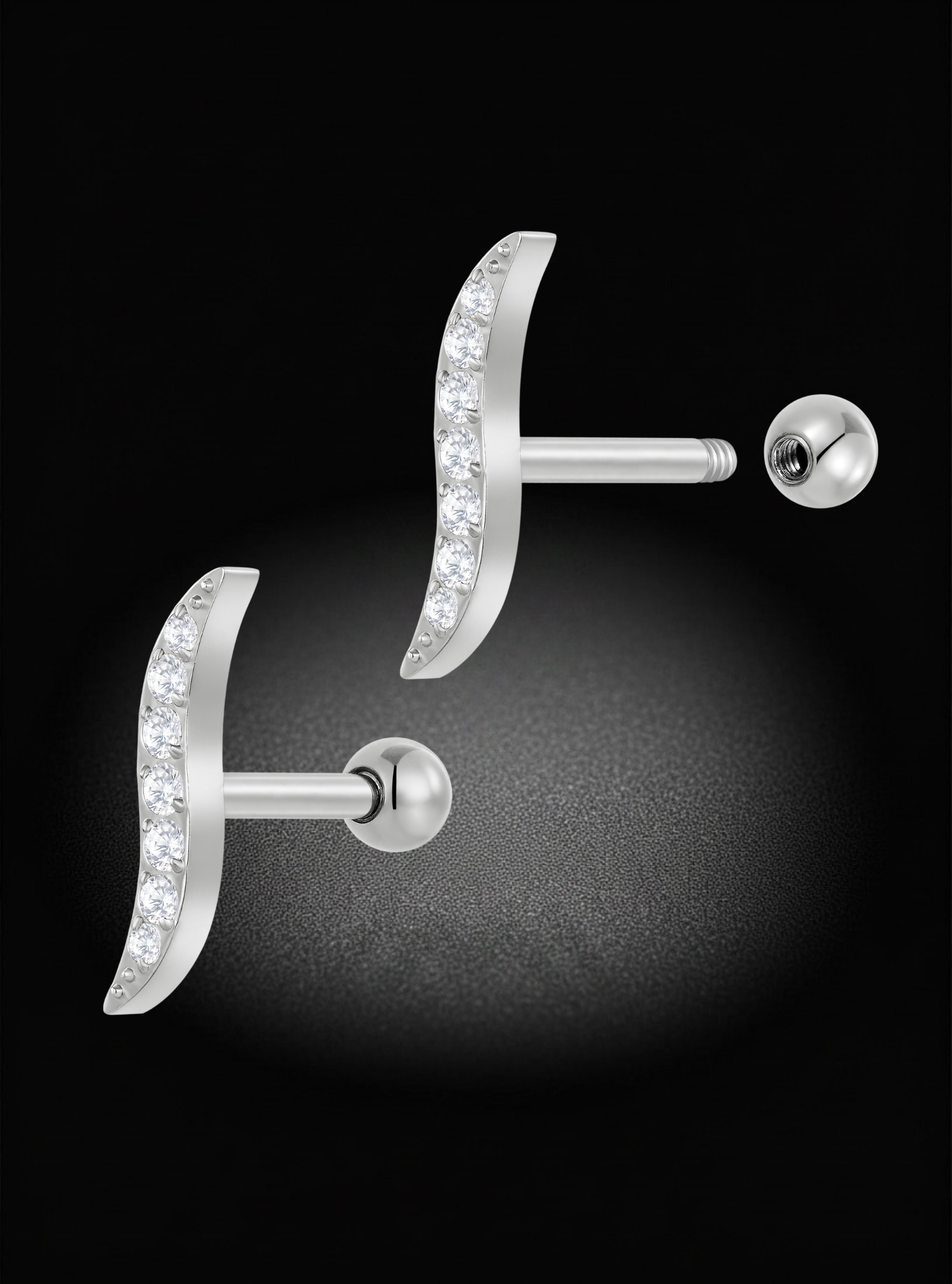 A pair of silver crescent moon stud earrings with clear pavé-set stones, shown against a dark black background with a soft spotlight effect.