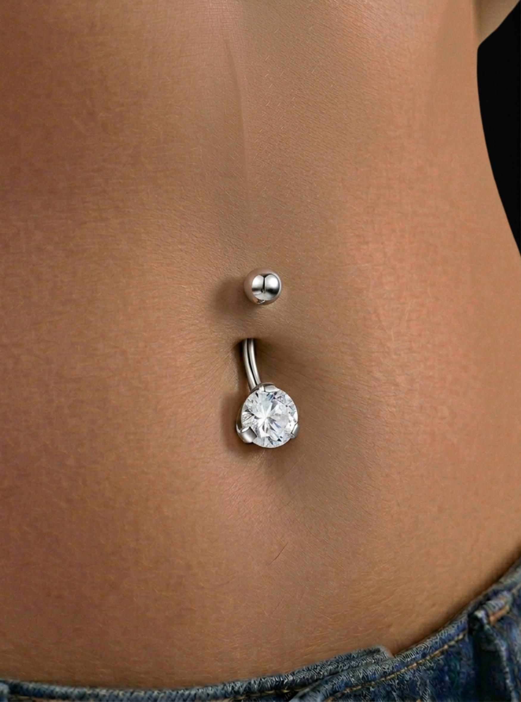 A close-up of a belly button piercing with a silver barbell, a small silver ball stud above it, and a larger round clear rhinestone pendant below, shown on a person's torso.