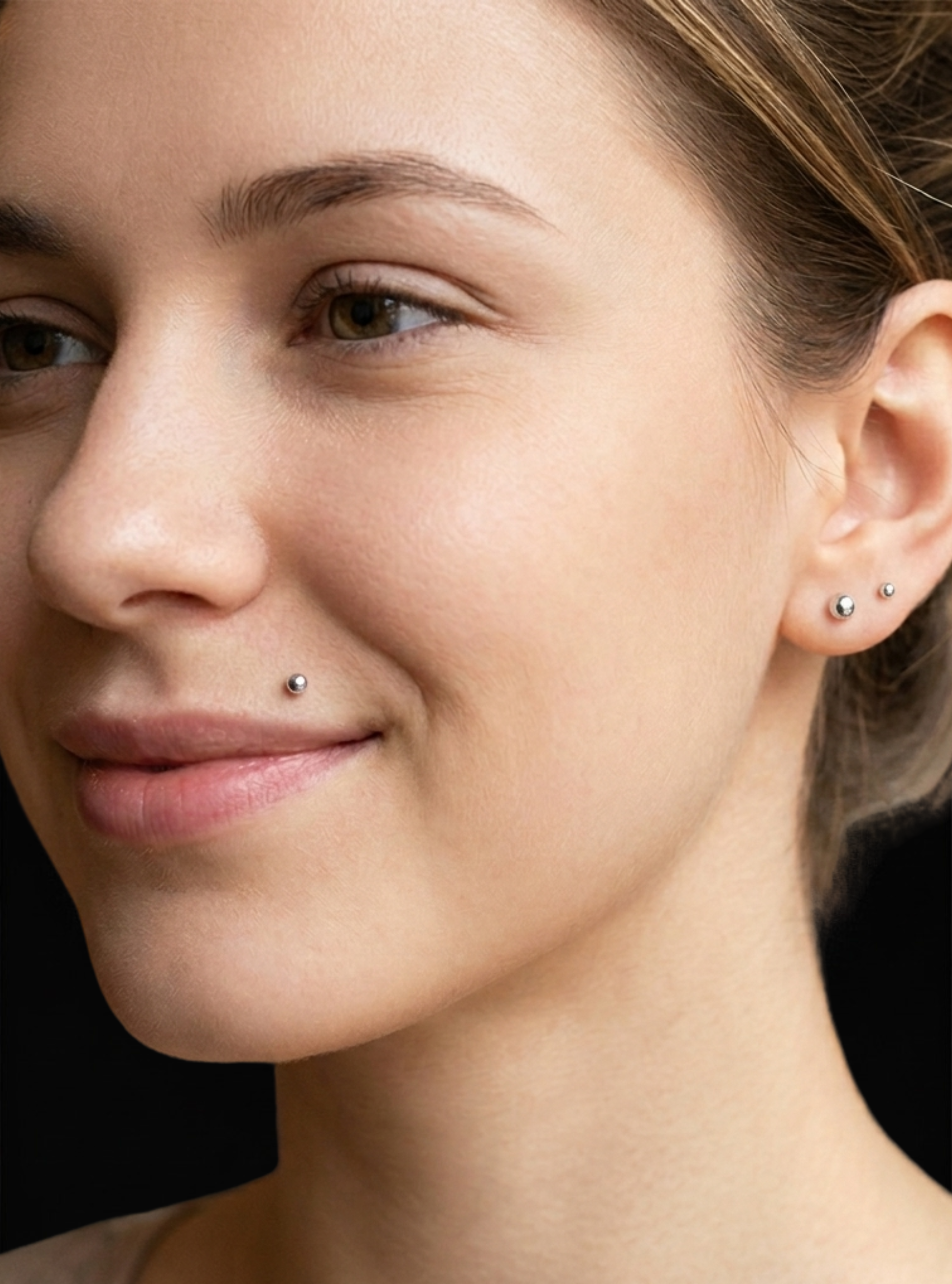 Close-up of a woman's face showing small silver stud earrings on her ears and a nose stud, against a dark background.