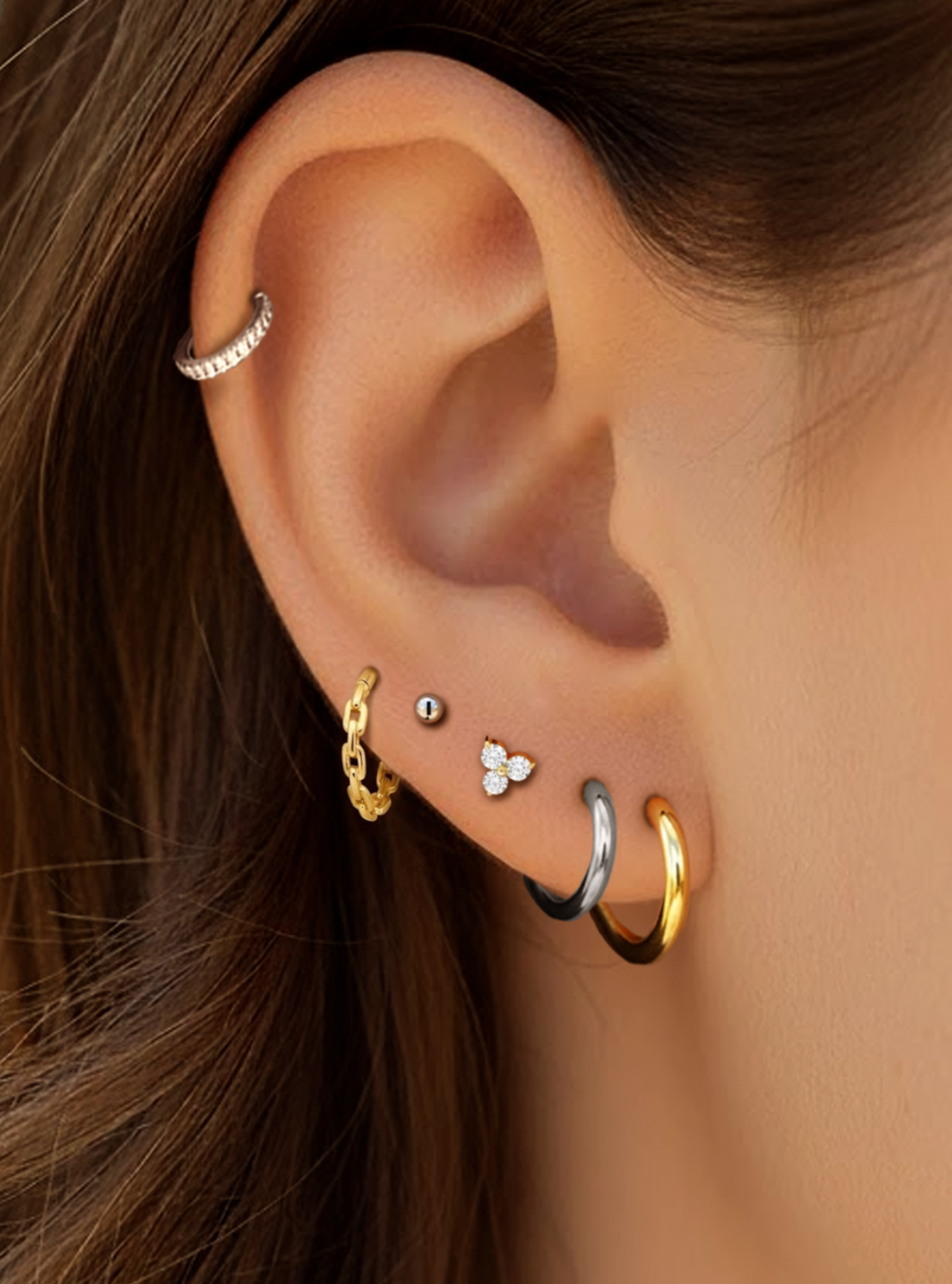 Close-up of a person's ear showcasing multiple stacked earrings, including a silver cartilage hoop, a gold huggie, a silver and black hoop, a gold hoop, a small silver stud, and a tiny white flower stud.